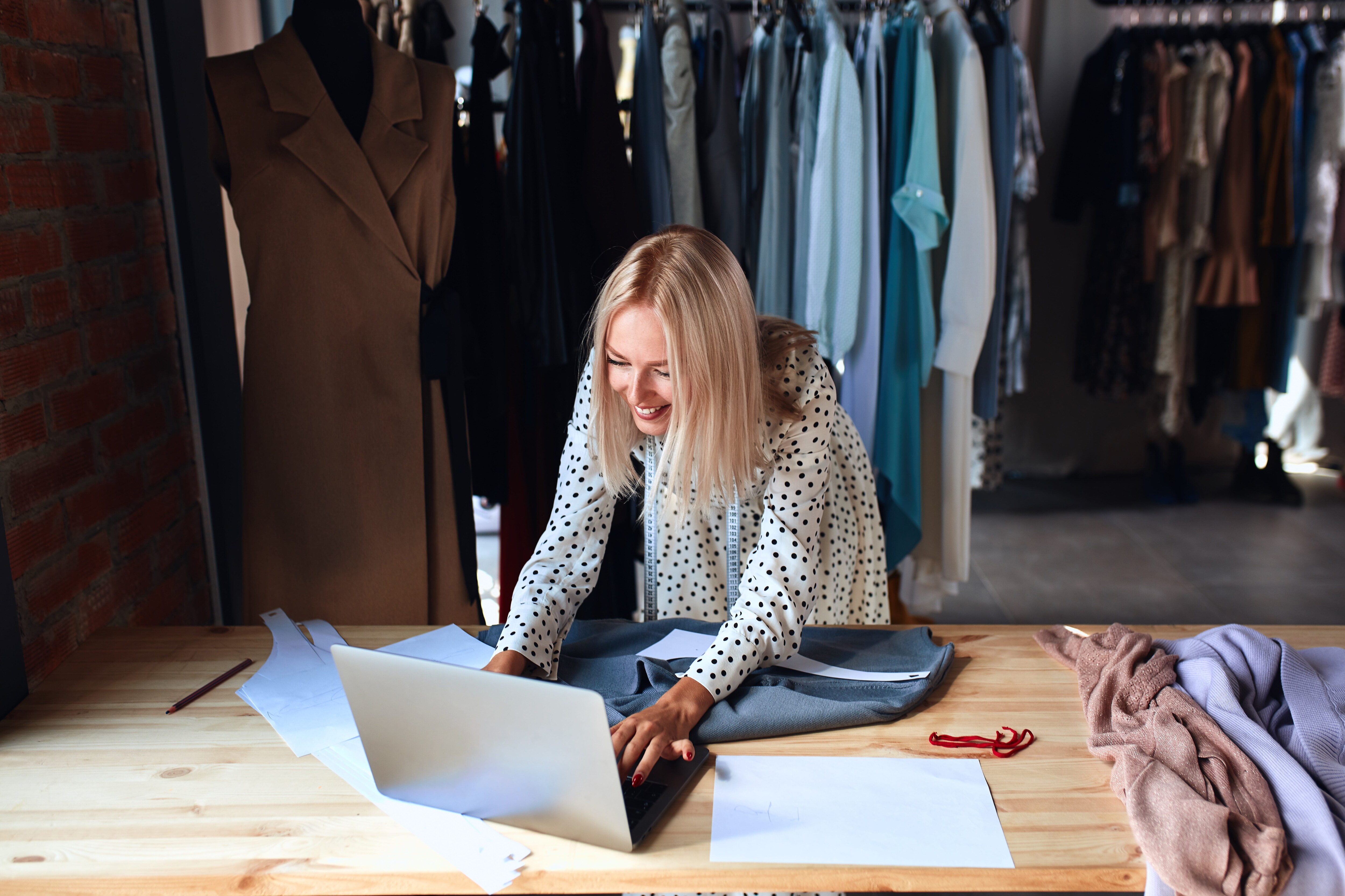 Designer working on a laptop with clothing racks behind her.jpeg
